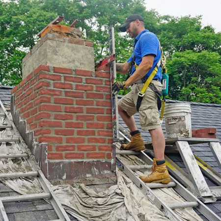 A chimney mason installing the final bricks on a chimney while safely working with harness equipment on a steep roof.